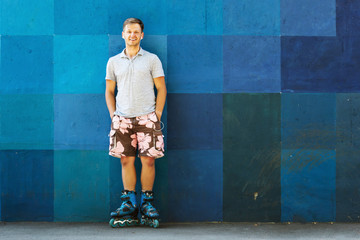 Happy handsome young roller in inline skates sitting against blue wall.