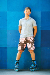 Happy handsome young roller in inline skates sitting against blue wall.