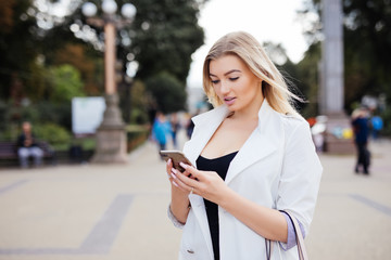 gorgeous beautiful young woman with blonde hair messaging on the smart-phone at the city street background.