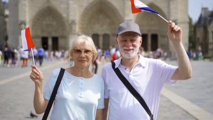 Happy cheerful family enjoying vacation. Seniors standing near Notre-Dame de Paris. Holding french flags in hands, waving it - Powered by Adobe