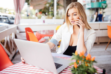 Fototapeta premium Young gorgeous woman having smart phone conversation while sitting in front of open laptop computer in cafe bar, attractive female calling with her cell telephone while she breakfast in coffee shop