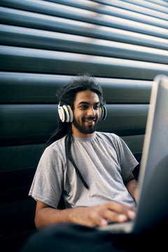 Indian young guy listening music trough the headphones and looking at laptop.