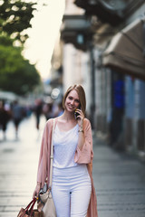 Fototapeta premium Beautiful and fashionable young woman with shopping bags standing on city street and looking at shopfront
