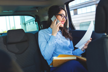 Beautiful young business woman sitting in the car looking at her planner and talking at phone.