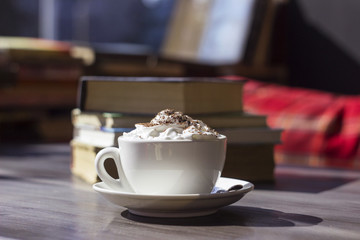 A white ceramic cup stands on the table in the background of books. A cup of coffee with foam and chocolate. A cafe