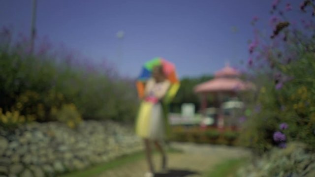 Happy sexy young woman with colored rainbow umbrella winks at camera, walks on footpath in flower garden. Pretty dreaming cute girl in funny lace dress is spinning, falling in love. Slow motion