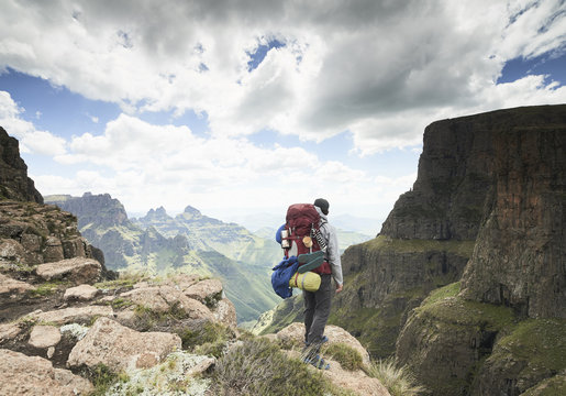A male hiker with his backpack standing at a mountain cliff edge over a mountainous green valley with clouds.
