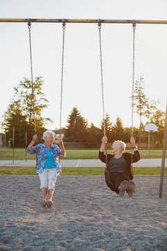 Two happy, senior caucasian friends swinging together on swingset at sunset