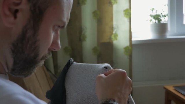 Brutal bearded man sewing a hole in his clothes by himself close up