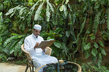 Arab muslim business man in white dress and hat sit on chair ,write down on notebook with cup of coffee and smart phone on glass table.