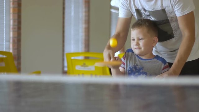 the father helps his son to play table tennis. the adult teaches the child how to hit the ball. the family is having fun. man accustoms the baby to sports games ping pong