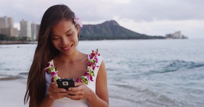 Happy young woman using smartphone at Waikiki Beach. Smiling female tourist is wearing orchid lei garland during vacation at island in Honolulu. She is text messaging using mobile phone on shore.