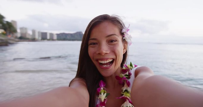 Cheerful young woman blowing kiss taking selfie video at Waikiki Beach. Happy beautiful female tourist is wearing orchid lei garland during vacation enjoying Honolulu, Oahu, Hawaii.
