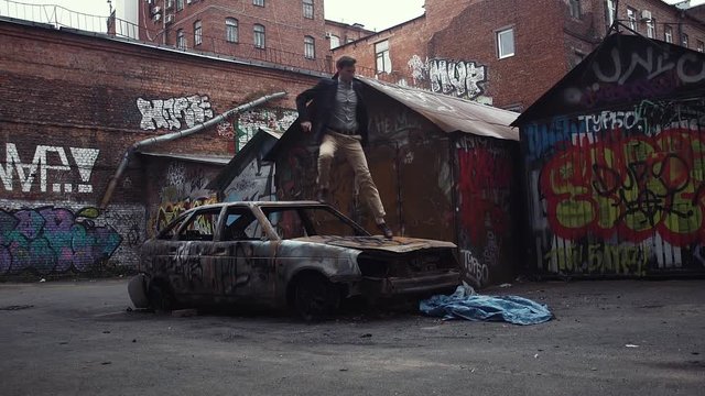 Young man running and jumping over roof of crashed burned car. Slow motion. Unidentifiable graffiti on the walls around. Urban lifestyle