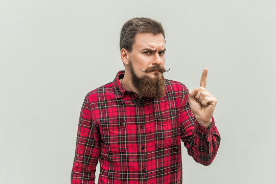 Warning sign. Young adult businessman with beard and handlebar mustache looking at camera with serious face and finger warning