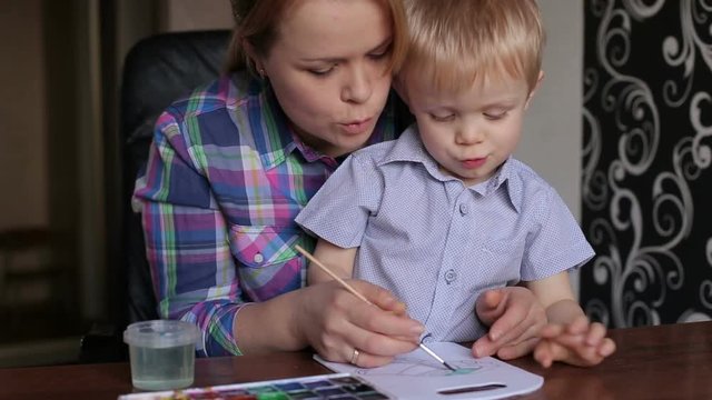 Young mother and her little son draw paints at home at a table.
