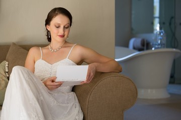 Beautiful bride reading wedding card while sitting on sofa