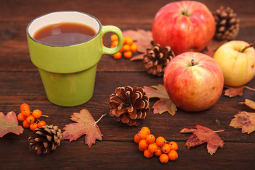 Autumn concept, fallen red-yellow leaves with apples, pine cones, rowan and a cup of tea on a wooden table. Thanksgiving Day.
