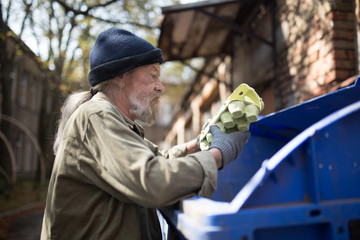 Homeles man searching food in thrash can. View of tramp holding packing for eggs.