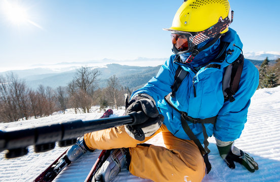 Close-up shot of a skier wearing colorful ski gear lying on the snow on top of a slope taking a selfie using camera on monopod selfie stick technology concept in the morning