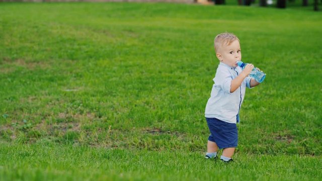 Funny kid 1 year is drinking water from a bottle. It stands on a green lawn, coolly staggers. Fun video with children