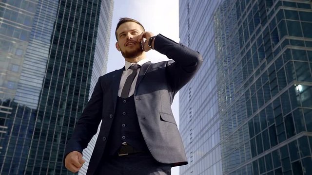 young manager is wearing three-piece suit is talking by phone, standing in background skyscrapers
