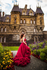 A beautiful woman, a queen in a burgundy lavish dress, walks along a flowering garden. Ancient castle on the background.
