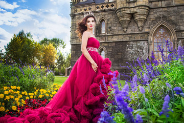 A beautiful woman, a queen in a burgundy lavish dress, walks along a flowering garden. Ancient castle on the background. © Надежда Манахова