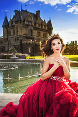 A beautiful woman, a princess in a red luxurious dress, sits by the fountain in a blooming garden. An ancient castle in the background. Medieval fantasy
