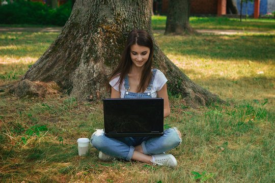 lovely young girl sits on a grass under a tree and using a lapto