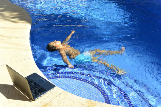 El ni&ntilde;o esta disfrutando el agua de la piscina mientras mirando su ordenador