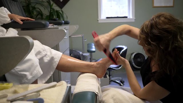 Pedicurist files her client's foot. Camera on slider tracking left to right.
