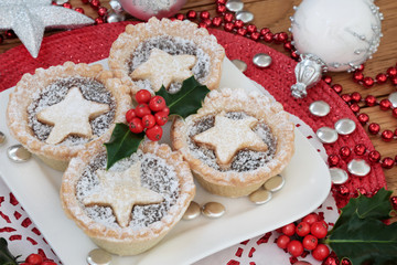 Christmas mince pie cakes on a plate with holly, bead, button  and bauble decorations on a white doilie on a red place mat.