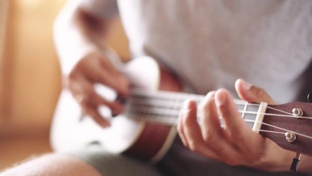 Crop shot of man holding and playing acoustic ukulele guitar sitting inside.
