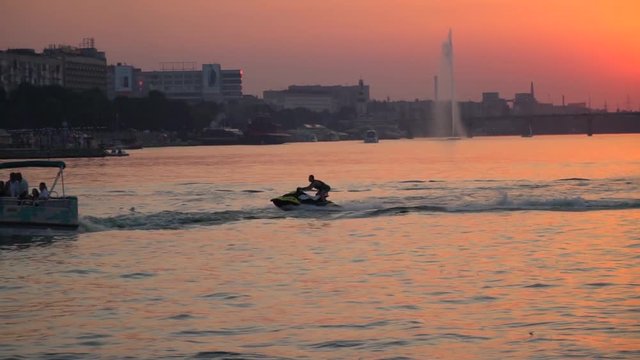 A male extreme on a hydrocycle rides near a slow boat against the background of the city embankment during sunset, slow motion