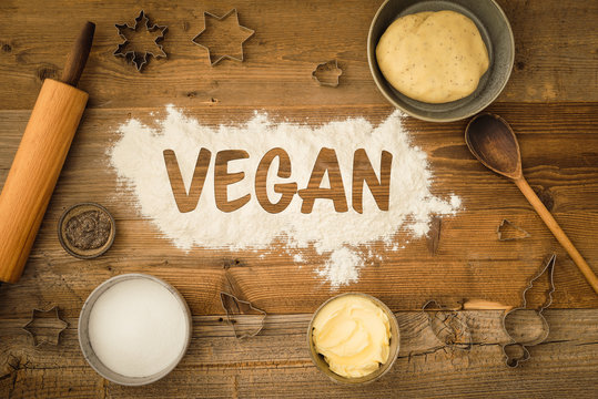 Flatlay collection of tools and ingredients for baking vegan Christmas cookies with margarine and chia seeds as egg replacement and the word vegan written in flour on a dark wooden table. Shot from