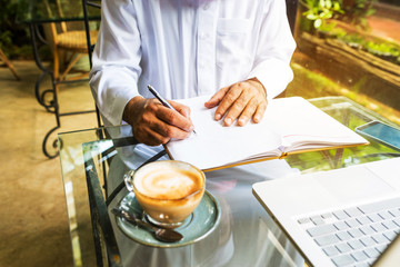 Arab muslim business man ware white traditional clothing and write on paper ,On table have laptop smartphone and cup of coffee
