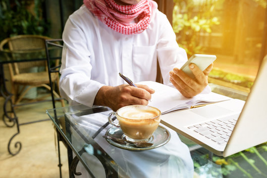 Arab Muslim Business Man Hand Writing On Paper Look At Smart Phone With Laptop On Coffee Glass Table