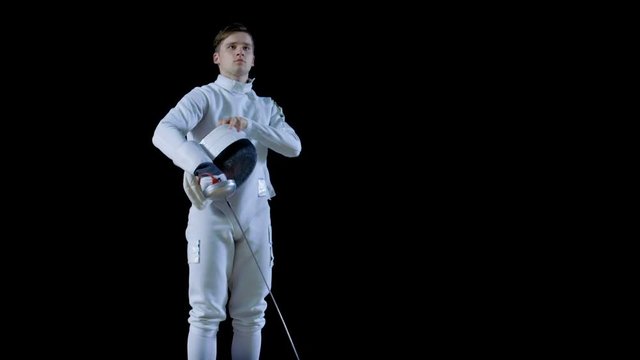 Portrait Shot of a Young Fencer Walking Into Focus while Holding His Protective Mask Under His Arm and Then Putting Him On and Making Lunge Attack. Shot Isolated on Black Background.