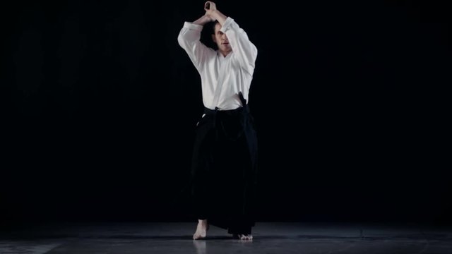 Aikido Master Wearing Traditional Samurai Hakama Clothes Takes His Japanese Sword out of Scabbard and Swings with It. He's in the Spotlight Darkness Surrounds Him. Shot Isolated on Black Background.