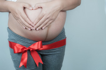 Closeup pregnant Woman holding her hands in a heart shape on belly with red ribbon, Happy pregnant woman Concept.