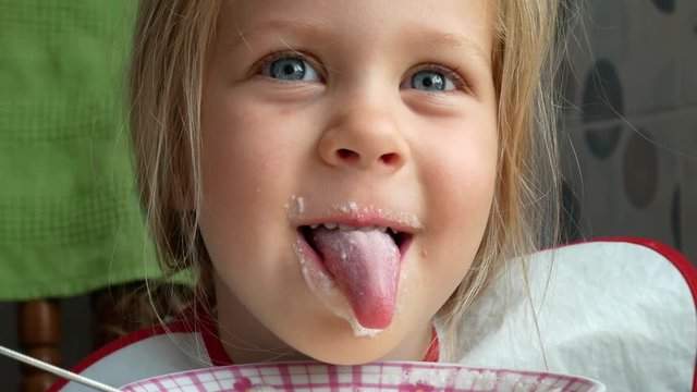 Little Beautiful Blonde Girl with Blond Hair and Blue Eyes Making a Mess After Eats Yogurt or Cottage Cheese Curd. Clumsy Dirty Girl Opens Mouth and Shows Tongue