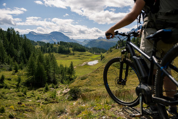 Naklejka na ściany i meble Young adult active man on mountain wearing bike helmet and backpack looking at scenic panorama holding electric bike in sunny summer day outdoor.