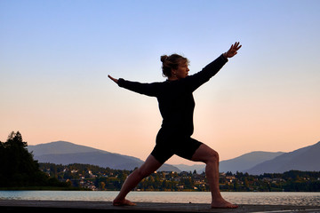 Fototapeta premium Mature woman in black clothes doing yoga on a wood jetty on Faaker See in Austria, early in the morning at sunrise