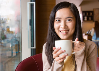 Asian woman drinking coffee in coffee shop next to window with blurry background