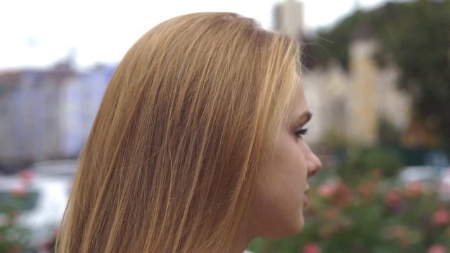 young blond girl with gray \ green eyes looking at the camera and smiling. a large female party in the background of a garden with roses. cloudy autumn or spring day. the girl is dressed in a warm