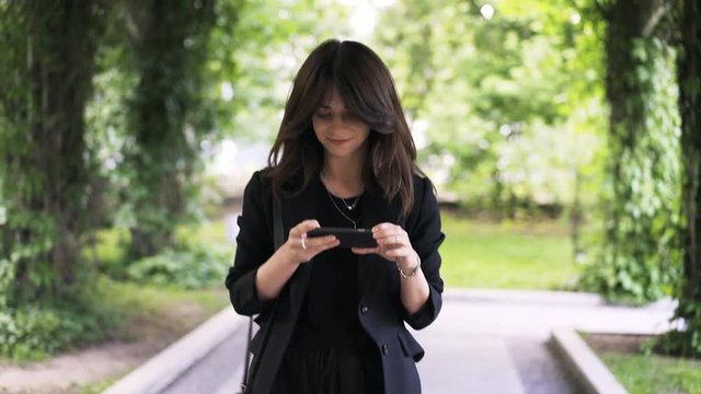 Attractive young woman wearing black is walking in a park and taking photos with her smartphone. Handheld real time medium shot