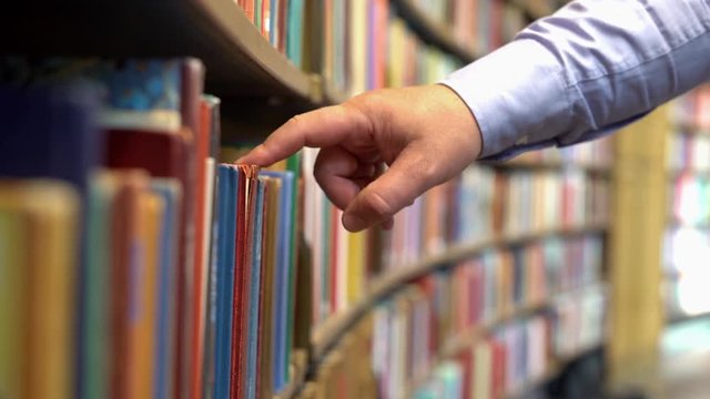 The hands of a man standing by a bookshelf in a library reading a book 