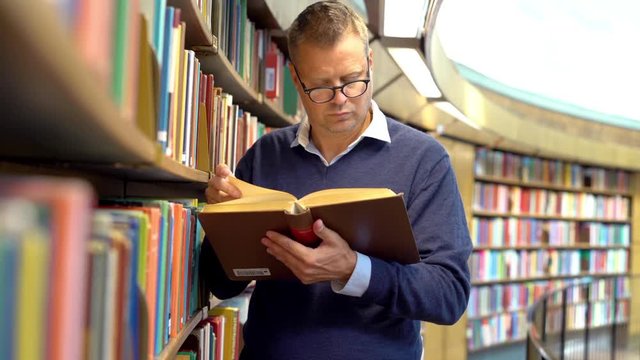 A man standing by a bookshelf in a library reading a book 