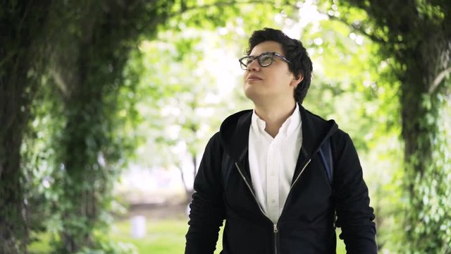 Handsome young man wearing glasses, a white shirt and a hoodie is walking in a park and having a good time. Handheld real time medium shot
